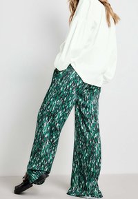 Zielone szerokie spodnie z wzorem, wykonane z gładkiego materiału, z abstrakcyjnym wzorem w odcieniach zieleni, czerni i bieli. Widać czarne obuwie.