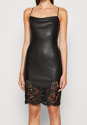 Robe noire moulante sans manches avec fines bretelles et détails en dentelle florale ajourée le long de l'ourlet, portée par une personne debout.