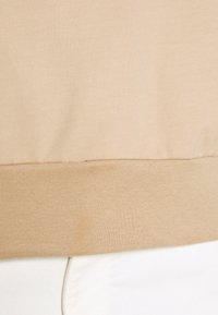 Ourlet de sweat-shirt beige avec bande côtelée sur pantalon blanc, montrant la texture du tissu et les détails des coutures.