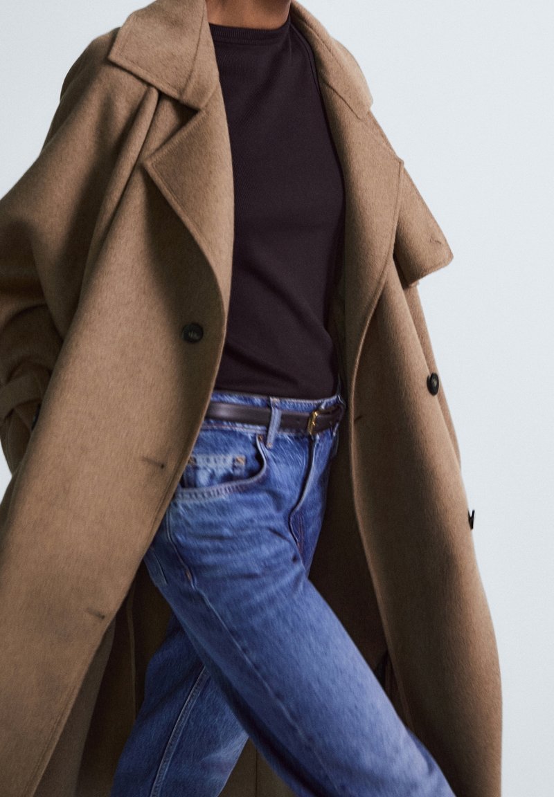 Personne portant un manteau en laine beige sur une chemise noire et un jean en denim bleu avec une ceinture marron, vue des épaules aux cuisses.