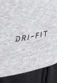 Der graue DRI-FIT-Stoff weist eine strukturierte Webart auf, mit einem genähten Saum und kontrastierendem schwarzem Stoff darunter, was ein sportliches Erscheinungsbild verleiht.