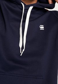 Felpa blu navy con cordini bianchi e piccolo logo bianco "G RAW" sul petto, che mostra la parte superiore del torso e la zona del collo.