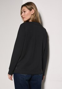 Maglietta nera a maniche lunghe con scollo rotondo, caratterizzata da una texture liscia e spacchi laterali. Indossata con jeans blu, presenta una silhouette casual.