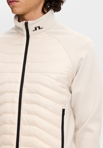 Mand iført let beige quiltet jakke med sort lynlås foran og lynlåslomme i siden, høj krave med sort logodetalje.