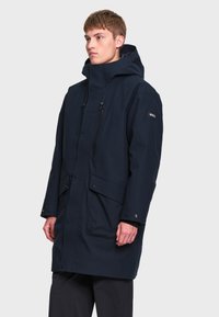 Jeune homme portant un long manteau d'hiver à capuche bleu marine foncé avec poches avant et fermeture éclair sur un fond clair uni.