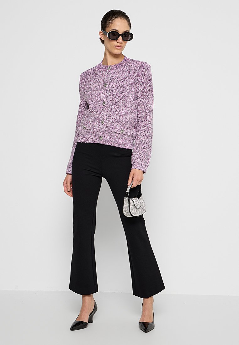 Lila strukturierte Strickjacke mit Rundhalsausschnitt und Knopfverschluss, kombiniert mit schwarzen Schlaghosen und einer kleinen silbernen Tasche. Model trägt Sonnenbrillen.