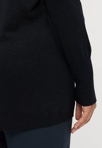 Pulover negru dintr-un material texturat, cu sclipiri subtile. Prezintă mansete și tiv cu dungi, fiind arătat aproape de un picior de pantaloni închisi la culoare.