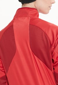Röd jacka med en kombination av släta och nätaktiga textilier. Designen inkluderar en hög krage och kontrasterande texturer på baksidan.