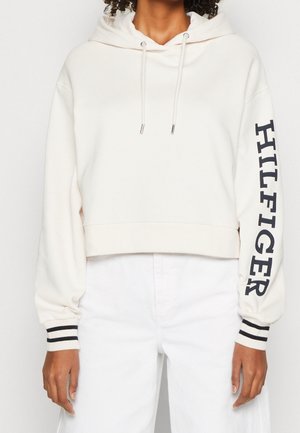 Kobieta w białej krótkiej bluzie z kapturem i sznurkami oraz napisem "HILFIGER" na prawym rękawie, w połączeniu z białymi spodniami z wysokim stanem.