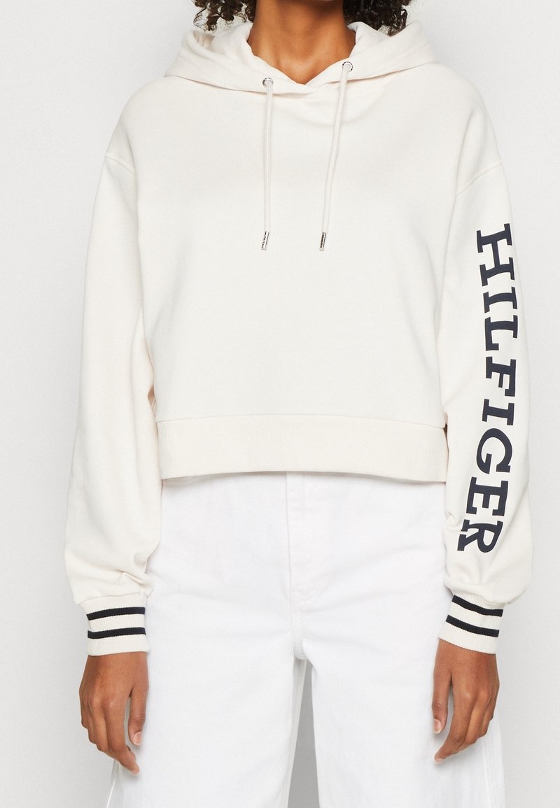 Femme portant un sweat à capuche court blanc avec cordons de serrage et le texte "HILFIGER" sur la manche droite, associé à un pantalon blanc taille haute.