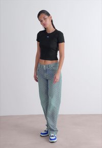 Crna uska crop majica, svijetloplave uzorkovane traperice i plavo-bijele tenisice. Model pozira protiv neutralne pozadine.