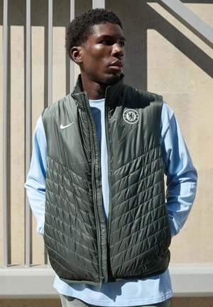 Green insulated vest with quilted design, Nike logo, and Chelsea FC badge. Worn over a light blue long-sleeve shirt. Casual fit and texture.