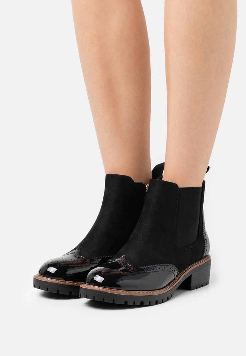 Bottines noires avec bouts brillants, talons épais et panneaux élastiques latéraux, portées sur des jambes nues sur un fond blanc.