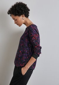 Zwart langemouwtop met een paars en rood abstract patroon, ronde halslijn en aangesloten manchetten, gecombineerd met zwarte broek.