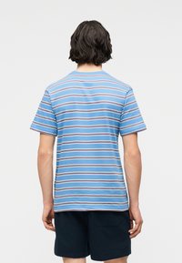 Άτομο με σκούρα μαλλιά μέχρι τους ώμους, φορεί μπλε κοντομάνικη ριγέ μπλούζα και μαύρο σορτς, με την πλάτη γυρισμένη, σε λευκό φόντο.