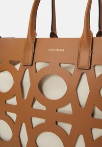 Sac fourre-tout en cuir tan avec des motifs géométriques découpés révélant un intérieur en toile crème. Doté de poignées en cuir marron et d'un logo doré.