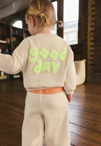 Sudadera beige claro con letras de espuma verdes que dicen "buen día" y detalles florales, combinada con pantalones a juego que tienen una cintura naranja.