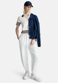 Žena stojí s rukama v kapsách bílých tepláků, má na sobě bílý polo triko s krátkým rukávem, tmavě modrou čepici, bílé tenisky a tmavě modrou bundu přehozenou přes rameno.