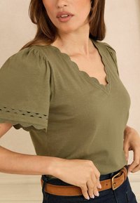 Žena v olivově zeleném vršku s vlnitým výstřihem do V a krátkými puff rukávy, s hnědým koženým páskem přes tmavé džíny.