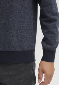 Navy blauwe sweatshirt met een gestructureerde, gespikkelde stof, gebreide manchetten en zoom, met een comfortabele pasvorm, getoond nabij de pols en hand.