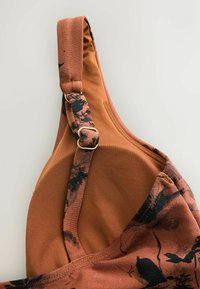 Bretelle de soutien-gorge en tissu marron avec imprimé floral noir et boucle dorée réglable sur fond clair uni.