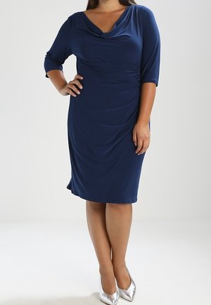 Kobieta w granatowej sukience do kolan z rękawami do łokci i srebrnymi, spiczastymi szpilkami, stojąca z jedną ręką na biodrze.