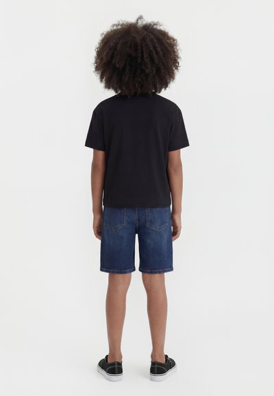 Παιδί με σγουρά μαλλιά στέκεται με την πλάτη, φοράει μαύρο μπλουζάκι, τζιν σορτς και μαύρα αθλητικά παπούτσια πάνω σε μονόχρωμο φόντο.