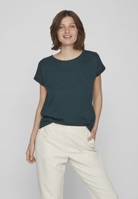 Dark teal short-sleeve t-shirt with a round neckline, paired with light beige, relaxed-fit trousers. Soft fabric, minimal design.