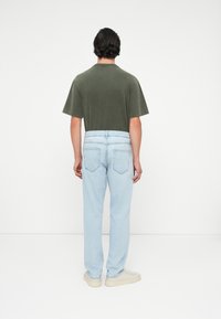 Grön bomullströja med korta ärmar, kombinerad med ljusblå denimjeans med bakfickor. Modellen står mot en neutral bakgrund.
