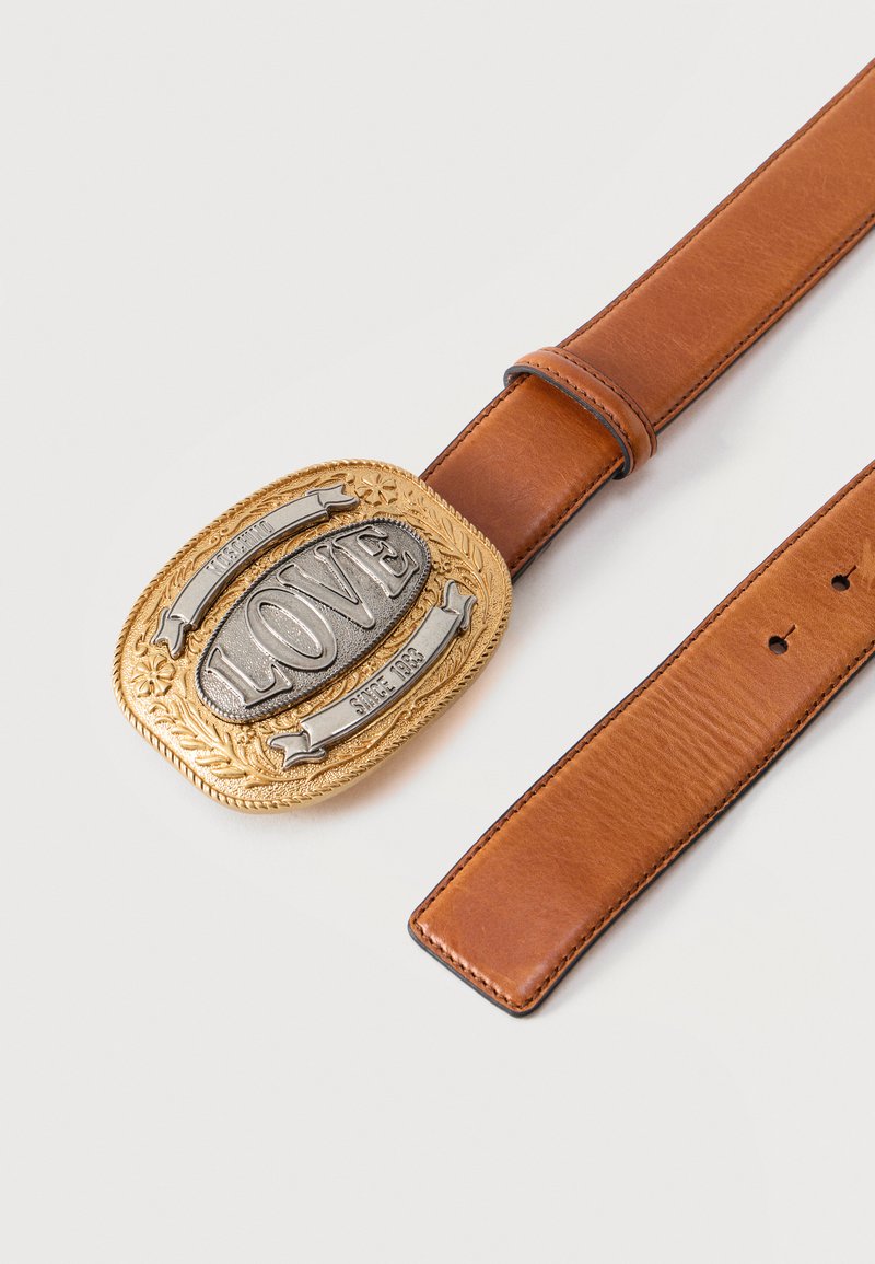 Ceinture en cuir marron avec une grande boucle ornée en or et argent portant l'inscription "LOVE" et "Depuis 1981" sur un fond blanc uni.