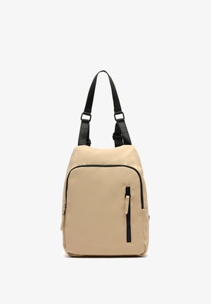 Zaino in nylon beige con accenti neri, tasca anteriore con zip e spallacci regolabili. Texture liscia, forma rettangolare e design minimalista.