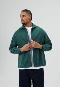 Grünes Hemd aus Wollmischung mit Knöpfen, Kragen, lockerem Schnitt, langen Ärmeln und dezenter Textur, über einem weißen T-Shirt getragen.