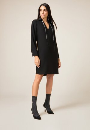 Schwarzes knielanges Kleid mit V-Ausschnitt, langen Ärmeln und silbernen Akzenten. Kombiniert mit funkelnden schwarzen Absätzen und strukturierten Socken.