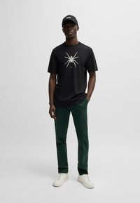 Tricou negru cu un grafic alb de păianjen, purtat cu pantaloni verzi închiși și adidași albi. Se poartă o șapcă neagră cu logo-ul "BOSS".