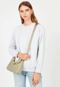 Sac en cuir beige doux avec un sommet noué, bandoulière réglable arborant un logo, associé à un sweat-shirt gris clair et un jean bleu.