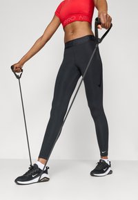 Černé legíny Nike Pro s vysokým pasem, hladkou texturou a perforovanými detaily. Kombinované s červeným zkráceným topem a černými sportovními botami.