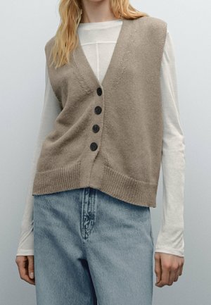 Femme portant un gilet en tricot beige boutonné par-dessus une chemise blanche à manches longues et un jean bleu clair taille haute sur fond blanc.