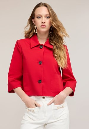 Jonge vrouw met lang blond haar, die grote gouden oorbellen draagt, een rood kort jasje met knopen en een witte broek met handen in de zakken.