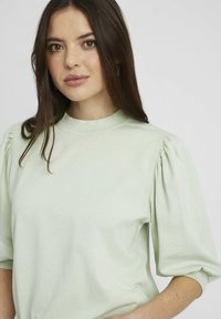 Sweatshirt vert clair avec un col montant, des manches bouffantes et un tissu lisse avec une texture subtile, créant une coupe relaxée.