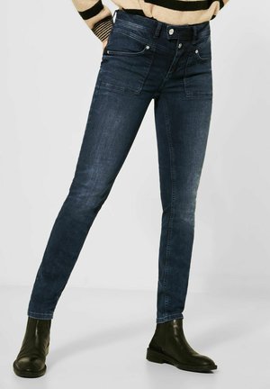 Femme portant un jean slim bleu foncé et des bottines noires, debout les jambes légèrement écartées devant un fond uni.