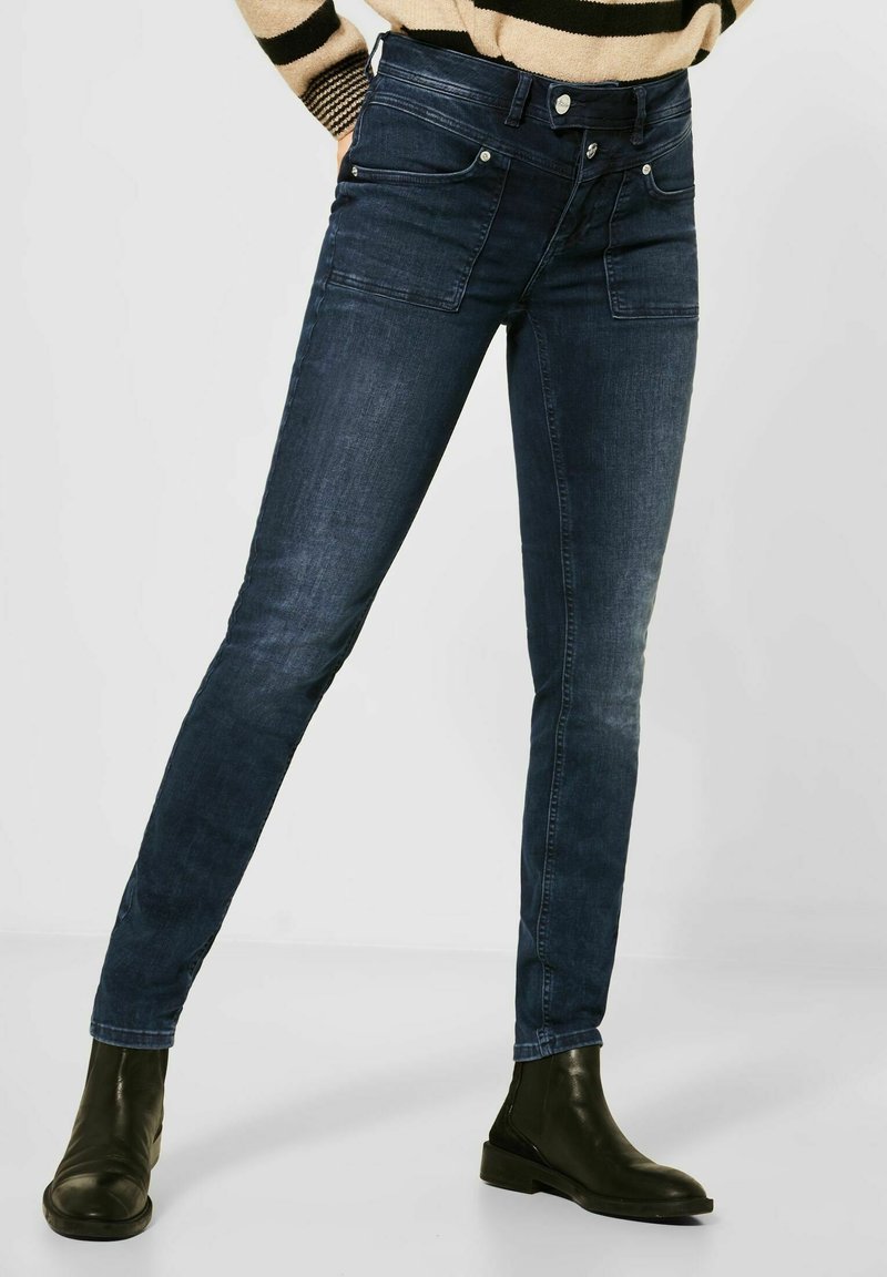 Femme portant un jean slim bleu foncé et des bottines noires, debout les jambes légèrement écartées devant un fond uni.