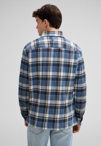 Jonge man met zijn rug naar de camera, gekleed in een blauw, wit en zwart geruit overhemd met lange mouwen en lichtblauwe jeans, tegen een effen grijze achtergrond.