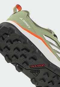 Grøn sportssko med grå og hvide detaljer, med en tekstureret sort gummisål mærket "TRAXION" og en trækflig med en orange accent.