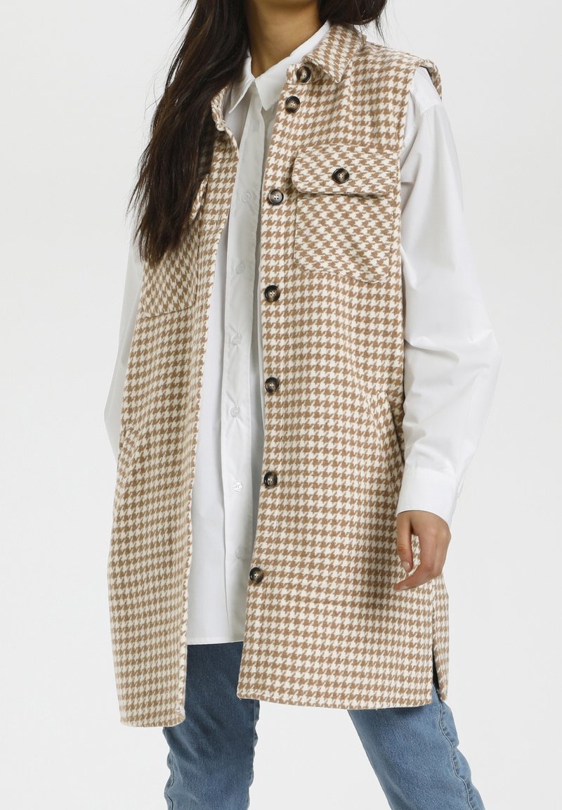 Gilet senza maniche con motivo a pied de poule nei toni beige e marrone, dotato di colletto, quattro tasche frontali e bottoni neri, indossato su una camicia bianca.