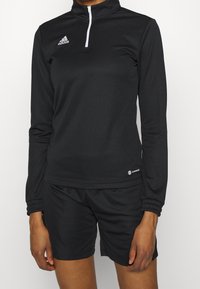 Personne portant un haut noir Adidas à manches longues avec fermeture éclair au quart et un short de sport noir, debout avec les bras détendus devant un fond blanc uni.