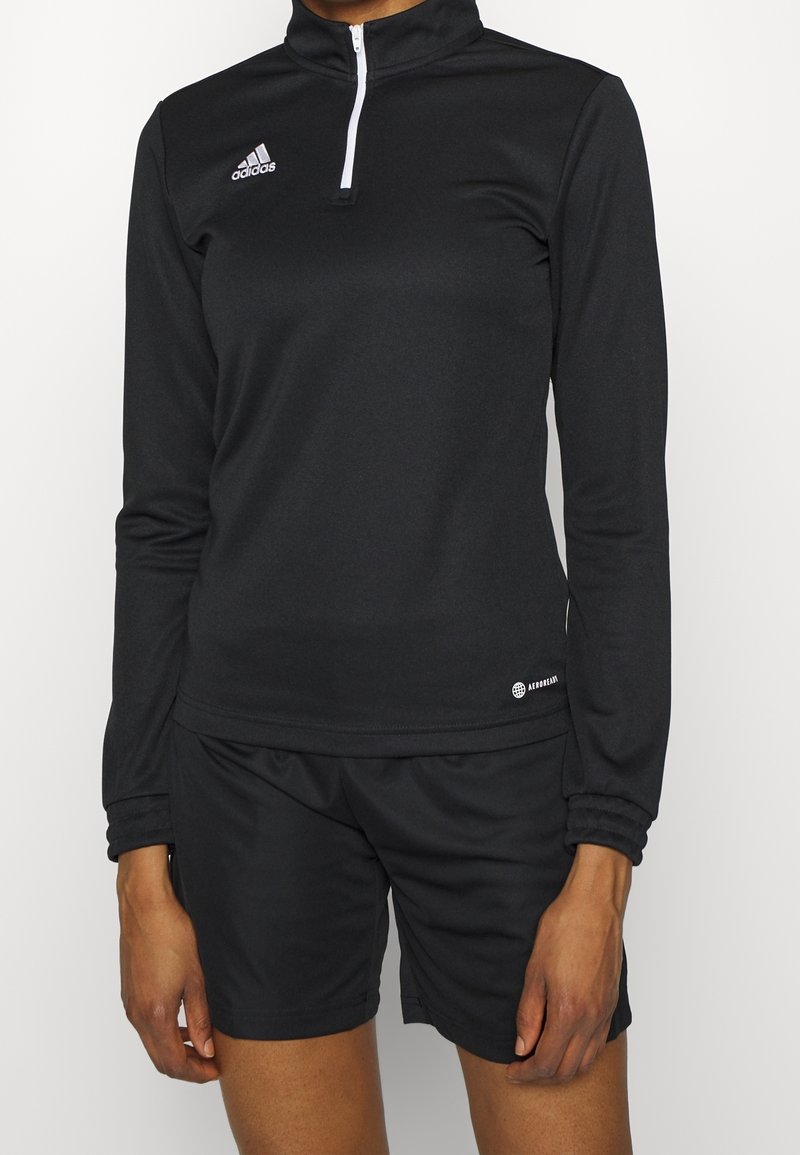 Personne portant un haut noir Adidas à manches longues avec fermeture éclair au quart et un short de sport noir, debout avec les bras détendus devant un fond blanc uni.