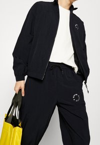 Marineblauwe windbreaker met hoge kraag en rits, gecombineerd met bijpassende joggers met een elastische tailleband en logodetails. Houdt een gele tas vast.