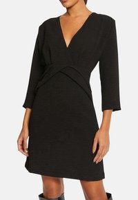 Robe noire avec un décolleté en V profond, des manches trois-quarts et un détail de taille noué. Texture douce et design à hauteur du genou.