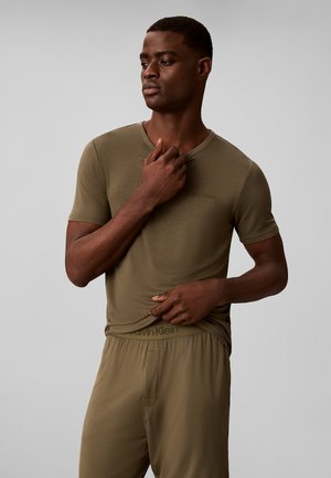 Hombre con camisa de manga corta Calvin Klein color verde oliva y pantalones a juego, ajustando el cuello de la camisa con una mano y sujetando el dobladillo con la otra.