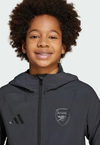 Veste zippée noire avec capuche, arborant un logo Arsenal sur la poitrine gauche et un tissu texturé. Comprend un logo Adidas sur la manche.